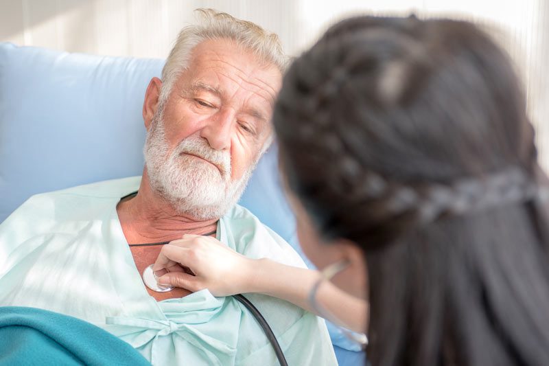Riverview Hospice and Palliative Care LLC | Nurse listening to an older man's heartbeat with a stethoscope as he lies in bed